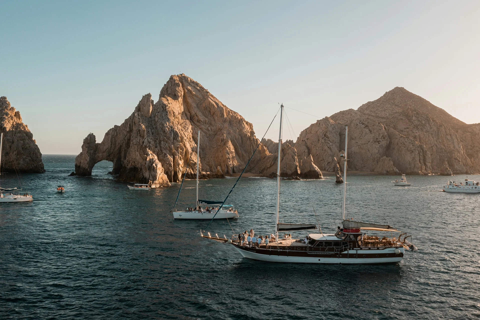 A dark-hulled wooden sailing ship anchored in the bay off El Arco at Cabo San Lucas, lit by the setting sun